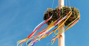 Uma grande fogueira acesa à noite, com pessoas a dançar ao redor com fitas e flores.